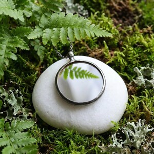 May include: A circular pendant with a dark metal frame and a reflective surface that mirrors a green fern leaf. The pendant hangs from a thin dark chain and rests upon a smooth white stone, surrounded by green moss and fern foliage.