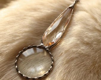 May include: A vintage-style magnifying glass with a clear, faceted crystal handle and a round, scalloped frame. The magnifying lens is clear, and the metal has a silver finish. The magnifying glass is resting on a light-colored, textured surface.