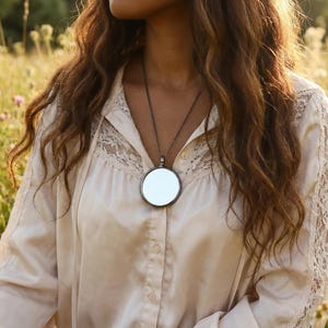 May include: A long, dark metal chain necklace featuring a large, circular, reflective mirror pendant. The necklace is worn over a cream-colored, long-sleeved button-down shirt that includes delicate lace paneling along the shoulders and chest.