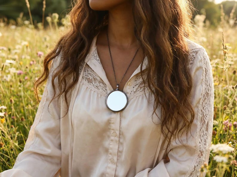 May include: A long, dark metal chain necklace featuring a large, circular, reflective mirror pendant. The necklace is worn over a cream-colored, long-sleeved button-down shirt that includes delicate lace paneling along the shoulders and chest.