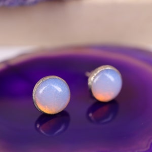 May include: Two opal stud earrings. Each earring features a round, iridescent opal stone set in a silver-colored bezel. The opals have a soft, pearly white and light blue color with hints of orange.