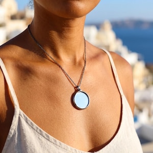 May include: A circular pendant necklace featuring a polished, reflective mirror surface encased in a dark, metallic frame. The pendant hangs from a thin, dark-toned metal chain that rests against the chest, providing a simple and clean aesthetic.