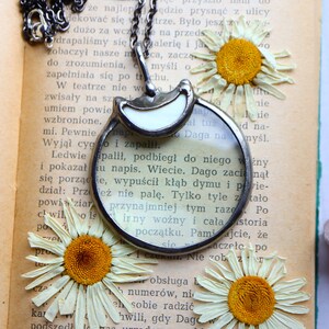 May include: A silver-toned magnifying glass pendant with a white accent, hanging on a chain. The pendant rests on an open book with dried daisies. A unique accessory for readers and vintage lovers.