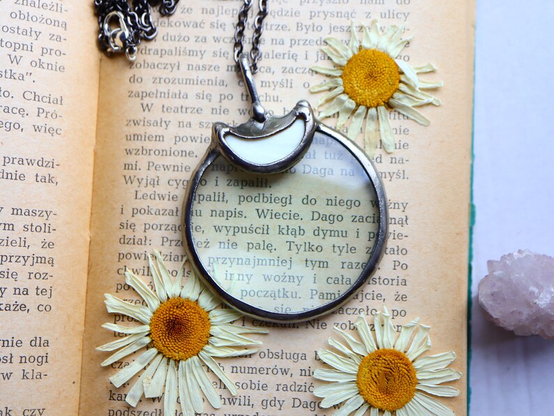 May include: A silver-toned magnifying glass pendant with a white accent, hanging on a chain. The pendant rests on an open book with dried daisies. A unique accessory for readers and vintage lovers.