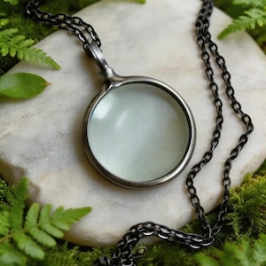 Könnte beinhalten: Eine silberfarbene Lupen-Anhänger-Halskette mit dunkler Kette. Die runde Lupe ist in einen kreisförmigen Rahmen gefasst. Die Halskette liegt auf einem weißen Stein, umgeben von grünem Moos und Farnen.