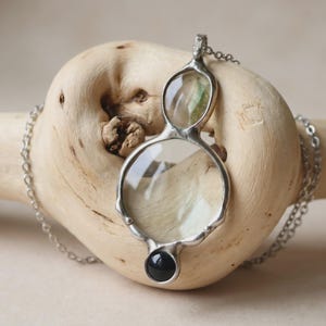 May include: A silver-toned pendant necklace featuring a vertical arrangement of two clear, rounded glass elements and a single small, opaque black circular stone at the base, all encased in a smooth, organic-shaped metal frame on a silver chain.