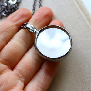 May include: A circular pendant necklace featuring a polished, reflective mirror surface encased in a dark, metallic frame. The pendant is attached to a dark, textured metal chain that extends upwards, showcasing a simple and functional design.