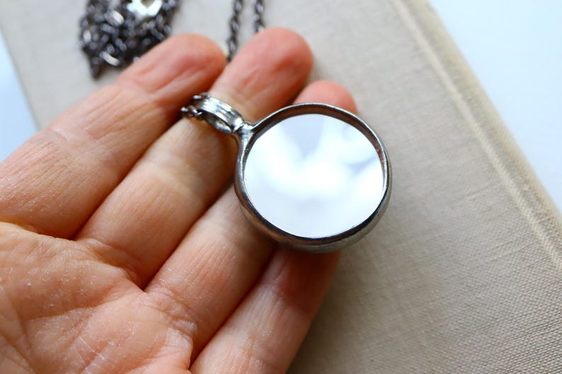 May include: A circular pendant necklace featuring a polished, reflective mirror surface encased in a dark, metallic frame. The pendant is attached to a dark, textured metal chain that extends upwards, showcasing a simple and functional design.