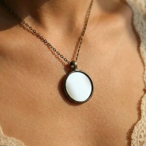 May include: A circular pendant necklace featuring a smooth, white, opaque center disc encased in a dark, antiqued bronze-toned metal frame. The pendant hangs from a matching thin, dark bronze-toned metal chain with small, uniform circular links.