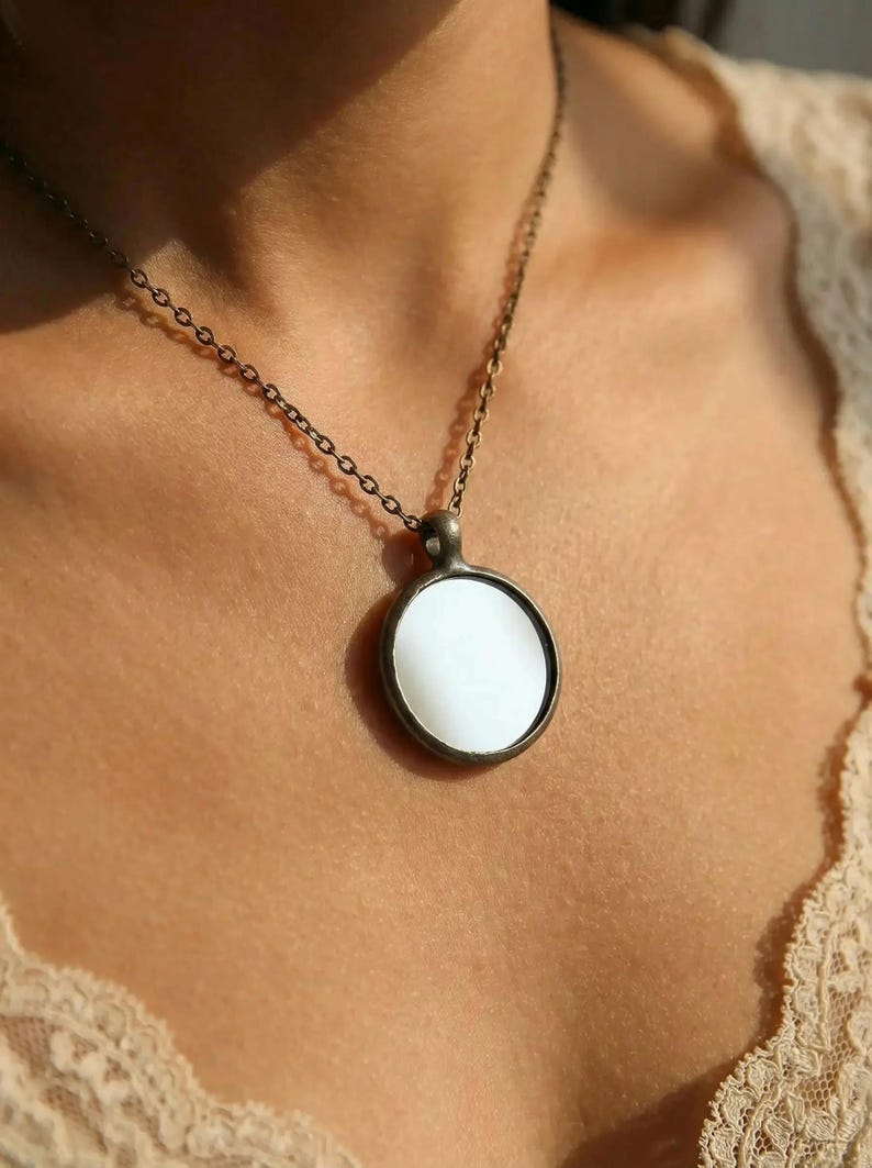 May include: A circular pendant necklace featuring a smooth, white, opaque center disc encased in a dark, antiqued bronze-toned metal frame. The pendant hangs from a matching thin, dark bronze-toned metal chain with small, uniform circular links.