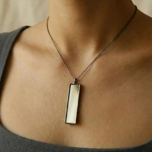 May include: A rectangular pendant necklace with a silver-colored frame and a white, reflective center. The pendant hangs from a delicate chain. The necklace is a simple, elegant piece of jewelry.
