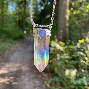 Könnte beinhalten: Eine silberne Halskette mit einem durchsichtigen, irisierenden Kristallanhänger. Der Kristall hat eine lange, spitze Form mit Regenbogenfarben. Der Anhänger ist mit einem silbernen Verschluss und einem kleinen, bläulichen Stein an der Kette befestigt. Der Hintergrund ist ein verschwommener Wald.