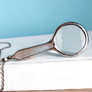 May include: A silver-toned magnifying glass pendant with a clear lens and a pale pink handle. The pendant hangs from a delicate silver chain. The magnifying glass is resting on a white surface against a light blue background.