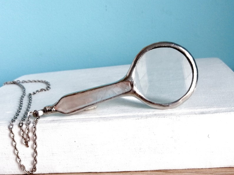 May include: A silver-toned magnifying glass pendant with a clear lens and a pale pink handle. The pendant hangs from a delicate silver chain. The magnifying glass is resting on a white surface against a light blue background.