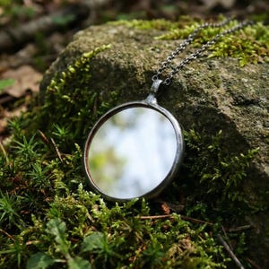 May include: A circular pendant necklace featuring a reflective mirror surface encased in a dark, oxidized metal frame. The pendant is attached to a thin, dark metal chain with small, uniform links, creating a simple and functional jewelry piece.