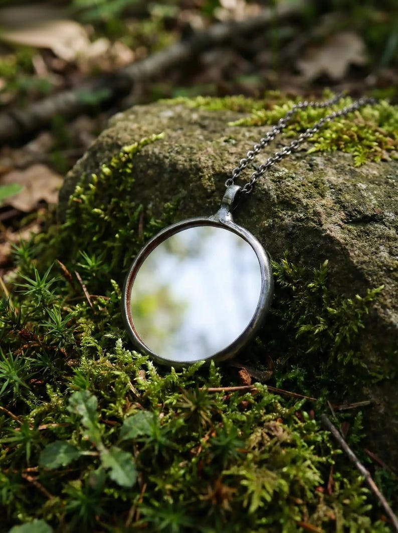 May include: A circular pendant necklace featuring a reflective mirror surface encased in a dark, oxidized metal frame. The pendant is attached to a thin, dark metal chain with small, uniform links, creating a simple and functional jewelry piece.