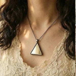 Op de afbeelding: Een ketting met een driehoekige hanger. De hanger heeft een crèmekleurig oppervlak en is omlijst door donker metaal. De ketting is ook van donker metaal. De ketting wordt gedragen over een crèmekleurige kanten top.