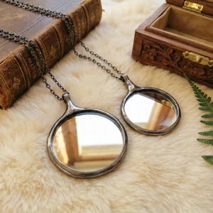 May include: Two circular mirror pendants on dark metal chains. Each pendant features a reflective glass surface encased in a dark, soldered metal frame with a small loop at the top for the chain attachment. The mirrors are of different sizes.
