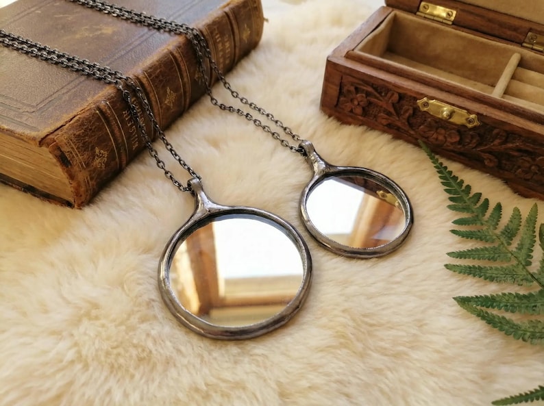 May include: Two circular mirror pendants on dark metal chains. Each pendant features a reflective glass surface encased in a dark, soldered metal frame with a small loop at the top for the chain attachment. The mirrors are of different sizes.