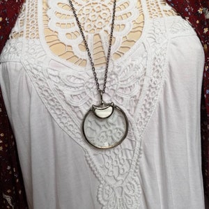 May include: A silver-toned magnifying glass pendant necklace. The pendant features a circular magnifying glass with a silver frame and a small, decorative element at the top. The necklace is displayed on a white top with lace detailing.