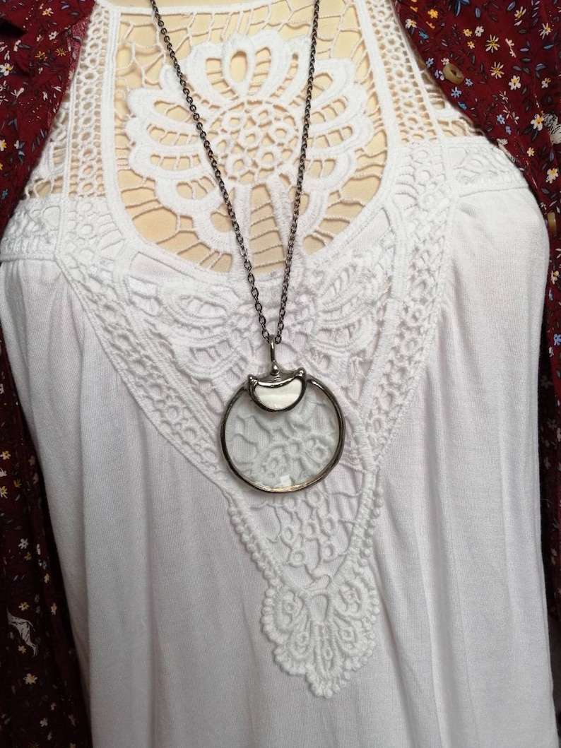 May include: A silver-toned magnifying glass pendant necklace. The pendant features a circular magnifying glass with a silver frame and a small, decorative element at the top. The necklace is displayed on a white top with lace detailing.