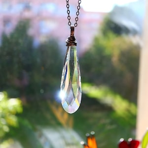May include: A clear, teardrop-shaped crystal pendant necklace. The crystal is faceted, catching the light and displaying a rainbow of colors. The chain is a dark, antique bronze color, and the pendant is suspended in front of a blurred green background.