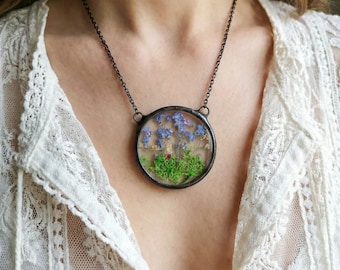 May include: A circular pendant necklace with a dark metal chain. The pendant features dried blue flowers, green moss, and a small red detail, encased in a clear glass frame. The necklace is worn over a white lace top.