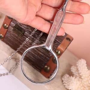 May include: A silver-toned magnifying glass pendant necklace with a clear lens and a handle inset with a pink stone. The pendant hangs on a delicate chain. The background includes a small wooden chest and coral.