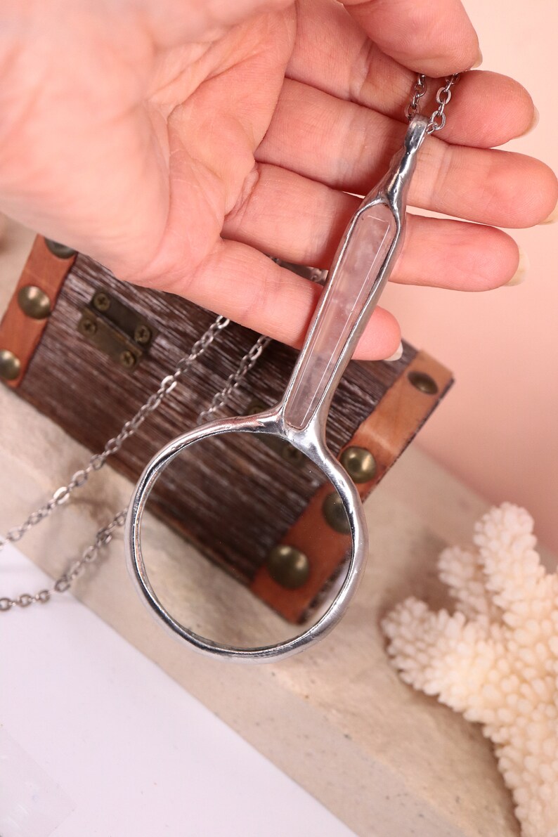 May include: A silver-toned magnifying glass pendant necklace with a clear lens and a handle inset with a pink stone. The pendant hangs on a delicate chain. The background includes a small wooden chest and coral.