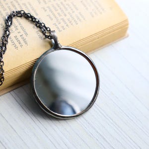 May include: A circular pendant necklace featuring a reflective mirror surface encased in a dark, metallic frame. The pendant is attached to a dark, textured metal chain, creating a simple and functional design that captures light and reflections.
