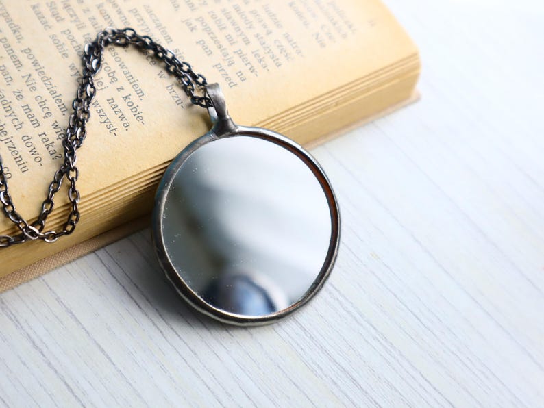 May include: A circular pendant necklace featuring a reflective mirror surface encased in a dark, metallic frame. The pendant is attached to a dark, textured metal chain, creating a simple and functional design that captures light and reflections.