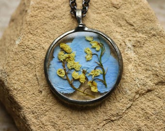 Puede incluir: Un collar con colgante circular que presenta un marco y una cadena de metal oscuro, conteniendo pétalos de hortensia azul claro prensados y pequeñas flores amarillas encapsuladas en resina transparente, creando un diseño botánico.
