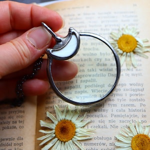 May include: A silver-toned magnifying glass pendant with a white crescent moon design. The pendant hangs on a chain and is held over an open book with dried daisies.