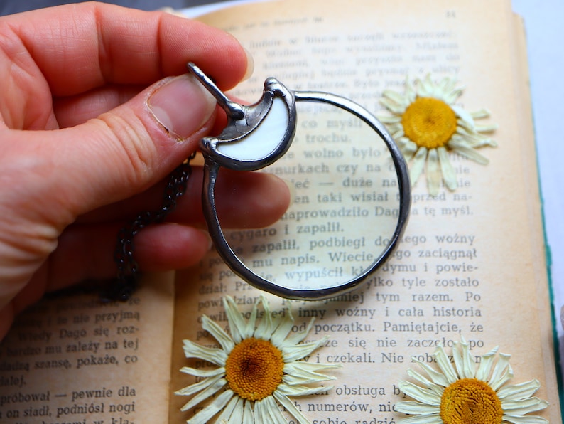 May include: A silver-toned magnifying glass pendant with a white crescent moon design. The pendant hangs on a chain and is held over an open book with dried daisies.