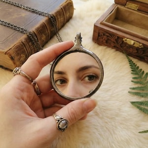 May include: A circular pendant mirror with a dark, textured metal frame and a thin chain. The mirror reflects a pair of eyes, showing a clear view of the iris and pupil. The pendant is held by fingers adorned with two silver rings featuring stones.