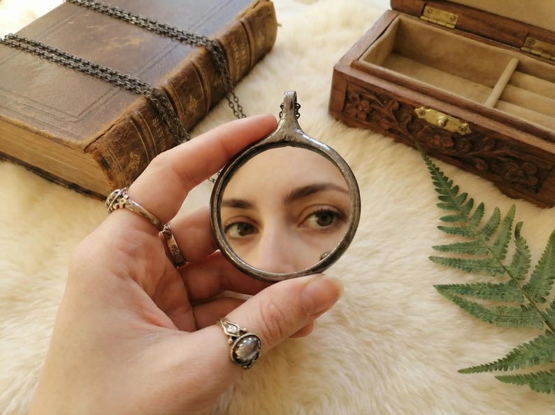 May include: A circular pendant mirror with a dark, textured metal frame and a thin chain. The mirror reflects a pair of eyes, showing a clear view of the iris and pupil. The pendant is held by fingers adorned with two silver rings featuring stones.