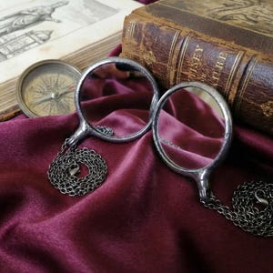 May include: Two circular silver-toned magnifying glasses with thin metal frames and attached dark metal link chains. Each magnifying glass features a clear glass lens set within a polished metallic rim, connected to a matching chain with a lobster clasp.