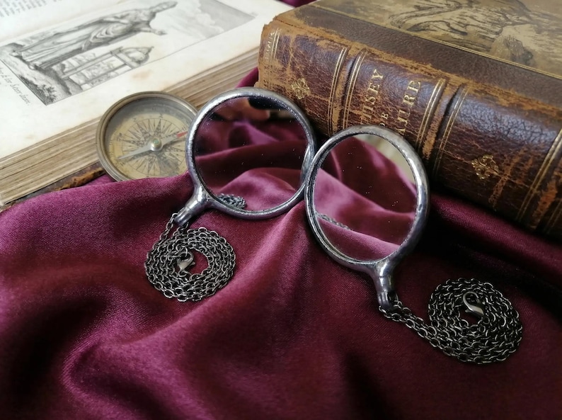 May include: Two circular silver-toned magnifying glasses with thin metal frames and attached dark metal link chains. Each magnifying glass features a clear glass lens set within a polished metallic rim, connected to a matching chain with a lobster clasp.