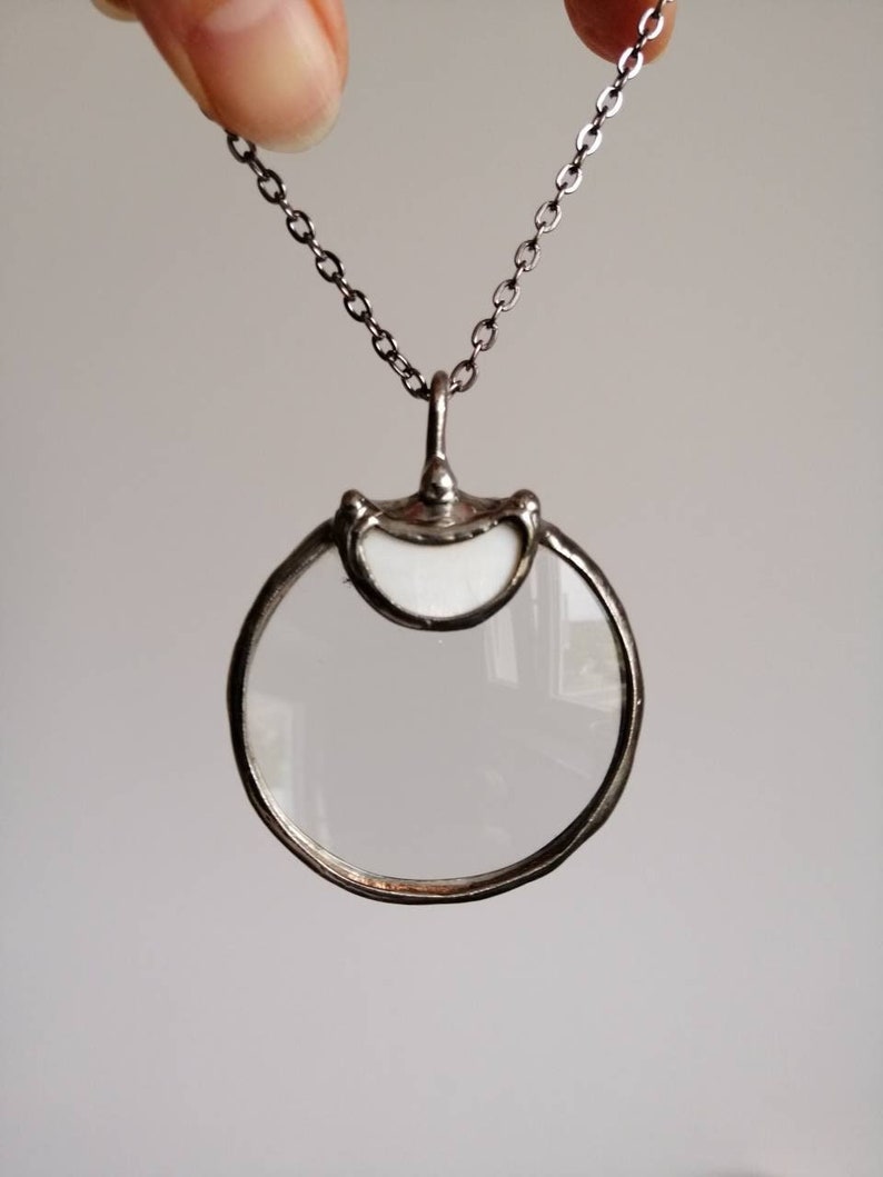 May include: A silver-toned pendant necklace featuring a circular magnifying glass. The pendant has a clear lens and a decorative frame. The chain is a delicate link style.
