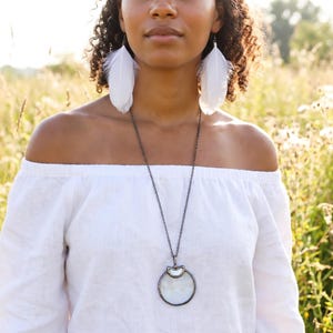 May include: A woman wearing white feather earrings and a long silver necklace with a circular pendant. She is wearing an off-the-shoulder white top. The background is a field of tall grass and wildflowers.