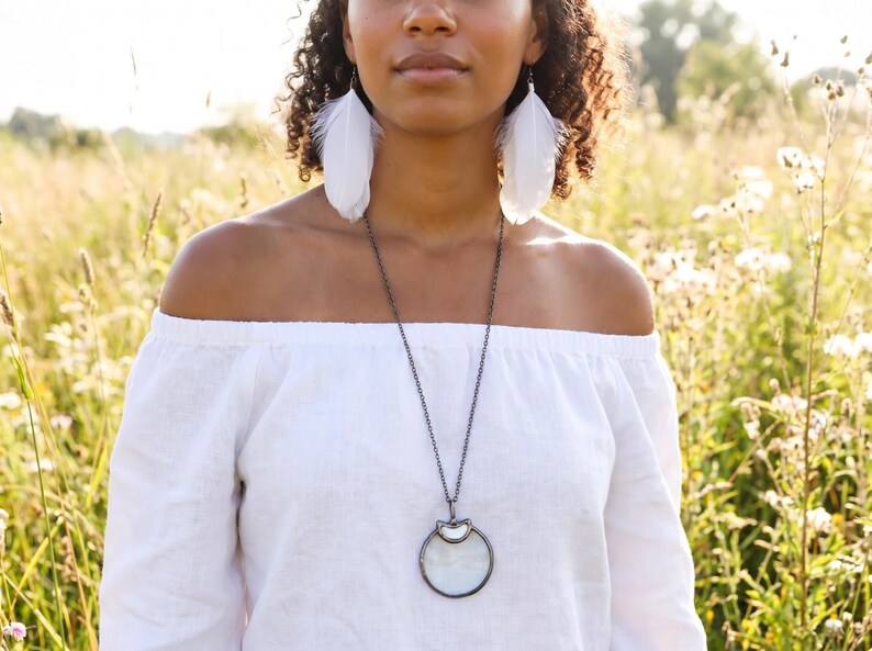 May include: A woman wearing white feather earrings and a long silver necklace with a circular pendant. She is wearing an off-the-shoulder white top. The background is a field of tall grass and wildflowers.