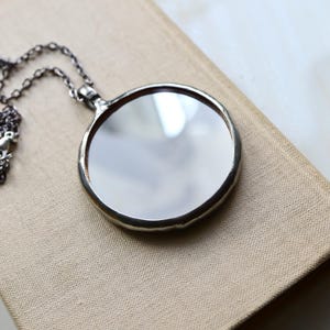May include: A circular pendant necklace featuring a reflective mirror surface encased in a smooth, dark metallic frame. The pendant is attached to a delicate, dark-toned metal chain with small, uniform links that extend from the top bail.