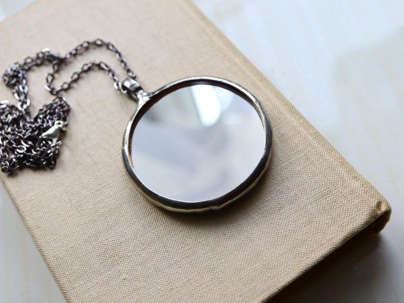 May include: A circular pendant necklace featuring a reflective mirror surface encased in a smooth, dark metallic frame. The pendant is attached to a delicate, dark-toned metal chain with small, uniform links that extend from the top bail.