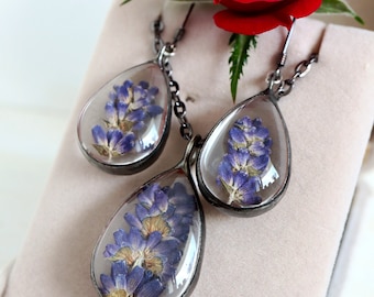 Op de afbeelding: Een sieradenset met een hanger en oorbellen, elk met een druppelvormige glazen behuizing. De behuizingen bevatten gedroogde blauwe bloemen. De set wordt gepresenteerd met een rode roos.