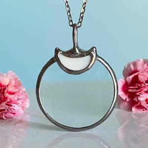 May include: A silver-toned pendant necklace featuring a circular magnifying glass design. The pendant has a white, curved top and a clear glass lens. The necklace is displayed against a light blue background with pink carnations on either side.