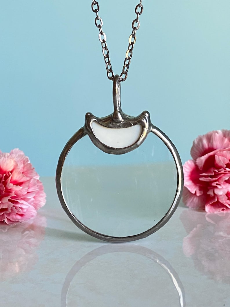 May include: A silver-toned pendant necklace featuring a circular magnifying glass design. The pendant has a white, curved top and a clear glass lens. The necklace is displayed against a light blue background with pink carnations on either side.