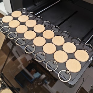 May include: A collection of twenty circular light-colored wooden keychains, each featuring a silver metal split ring, arranged in a grid pattern on a black rectangular 3D-printed tray with visible layer lines and the partial text TOOL.