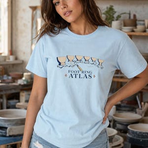 May include: A light blue short-sleeve t-shirt featuring a graphic illustration of five ceramic bowl bases in a row above a metal pottery tool. The text "FOOT RING ATLAS" is printed in dark blue letters below the illustration with small orange accents.