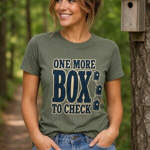 May include: An olive green short-sleeve t-shirt featuring the dark blue text ONE MORE BOX TO CHECK with three small birdhouse graphics arranged vertically on the right side. The shirt has a crew neckline and a standard fit for casual wear.