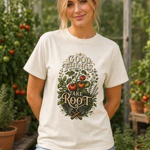 May include: A cream-colored short-sleeve t-shirt featuring a graphic of tomato plants, gardening trowels, and the text "GOOD THINGS TAKE ROOT" in a decorative font. The design includes red tomatoes, green leaves, and brown-handled tools.